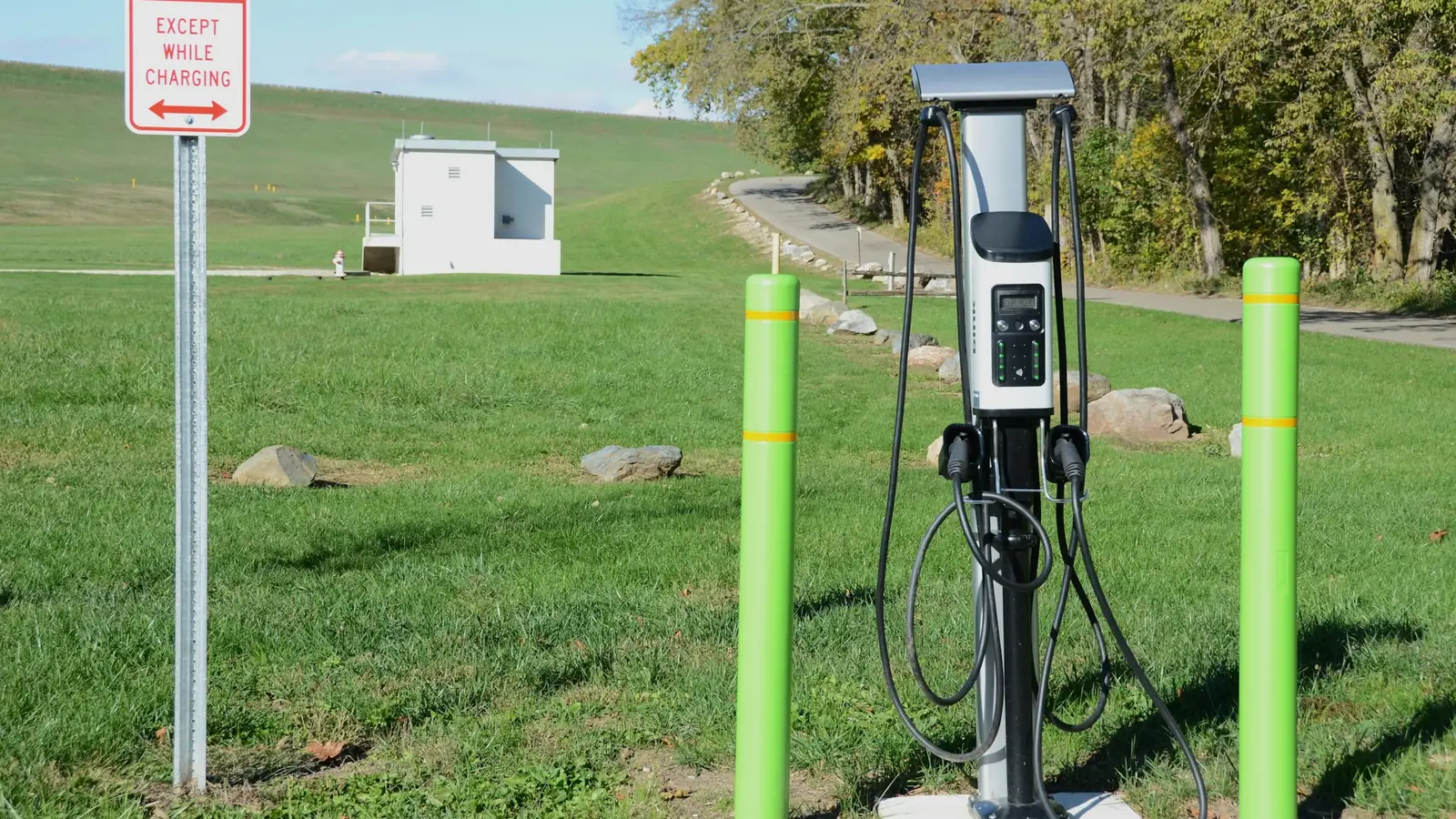 Public Level 2 EV charging station with two cables in a park, between two green bollards, next to a "no parking except while charging" sign