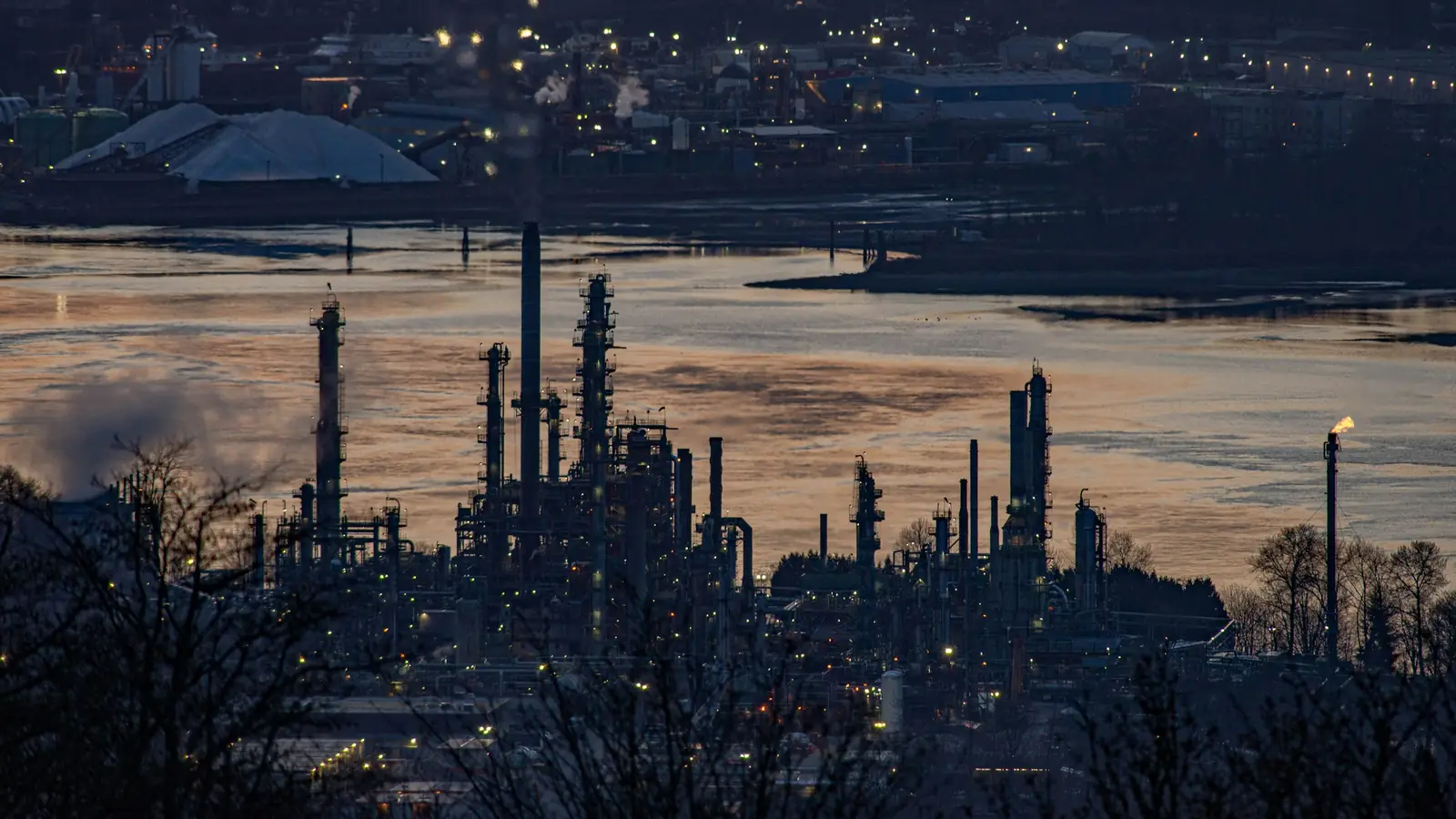 Oil refinery on the waterfront at dusk, with distillation columns and a lit flare stack, in Burnaby, British Columbia
